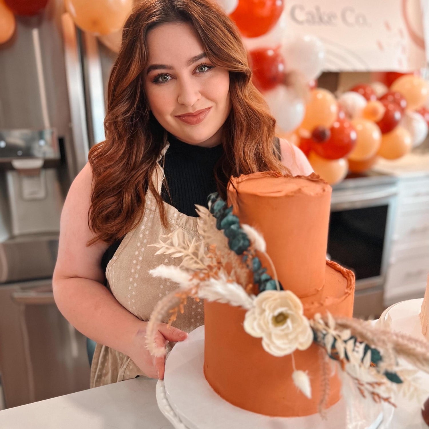 The owner of Lennon Cake Co smiles as she presents a beautiful two tiered cake with flowers.